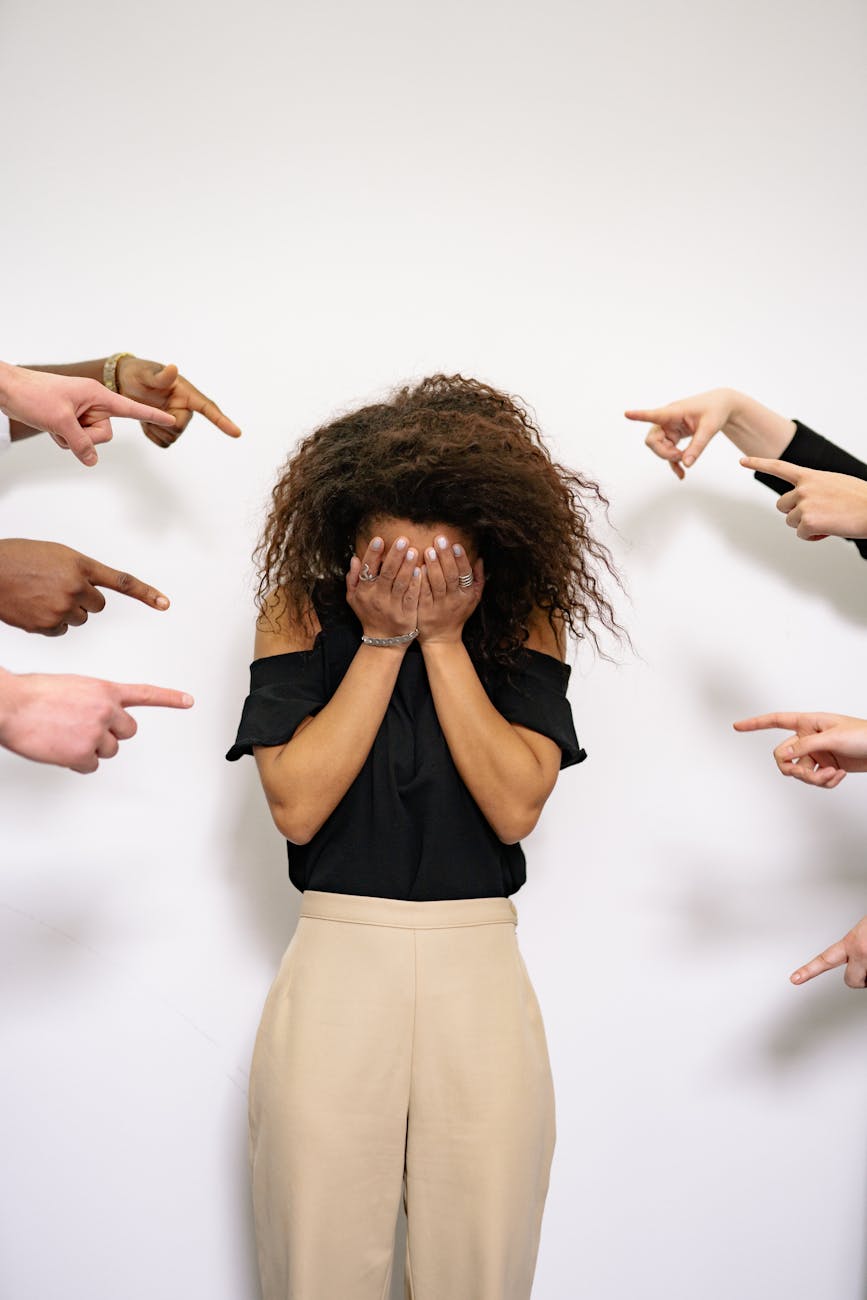 A brown woman with natural hair hiding her face in her hands while a bunch of hands point at her from various directions